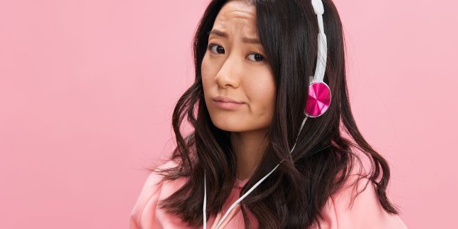 Upset confused Asian student young lady in pink hoodie sweatshirt with cute headphones looks at you posing isolated on over pink studio background. Good offer. Sound streaming platform ad concept